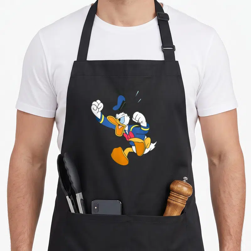 Anger duck apron - Cartoon Donald Duck with floating hat, blue suit, red bow tie, and clenched fists — shown on a black cotton canvas apron against a white background