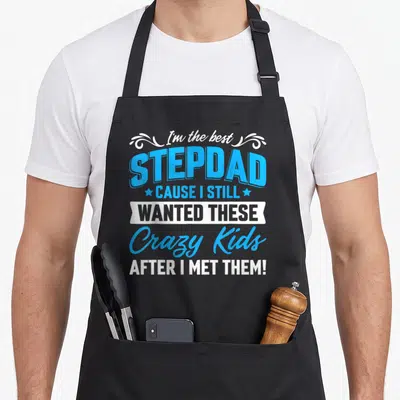 Stepdad family father day apron - Blue and white stacked text reading I'm the best STEPDAD CAUSE I STILL WANTED THESE — shown on a black cotton canvas apron against a white background