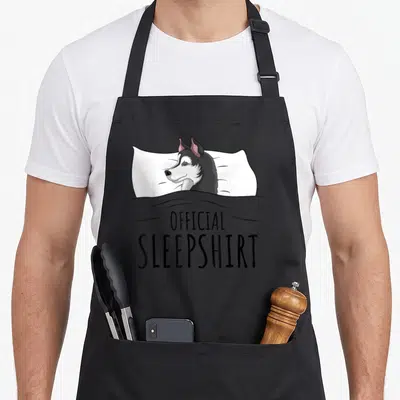 Sleepshirt dog apron - Cartoon husky sleeping under grey blanket with Official Sleepshirt text below — shown on a black cotton canvas apron against a white background