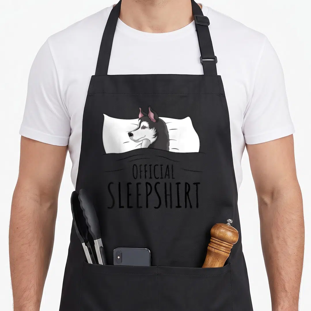 Sleepshirt dog apron - Cartoon husky sleeping under grey blanket with Official Sleepshirt text below — shown on a black cotton canvas apron against a white background