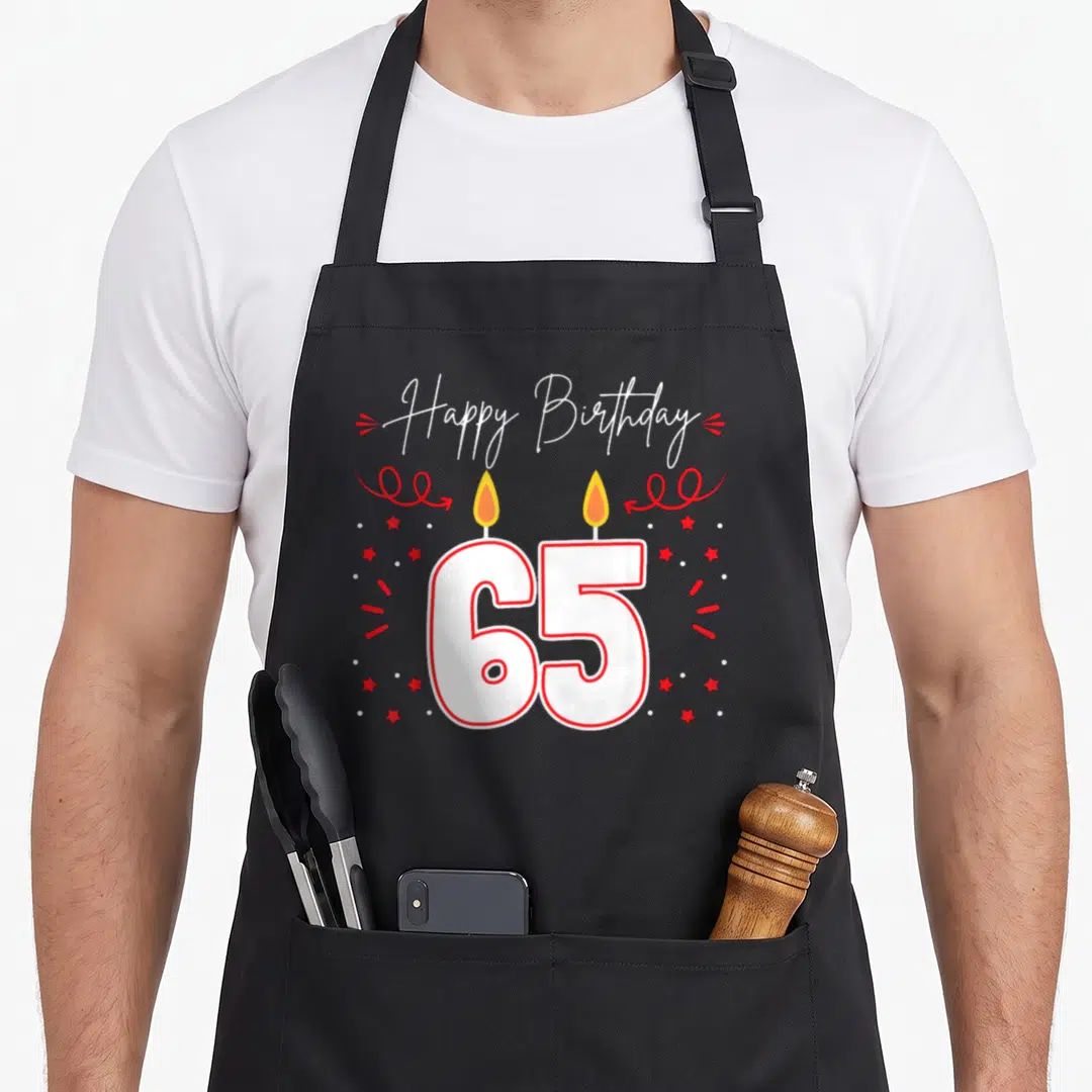Candlelight birthday apron - Happy Birthday 65 text with two yellow candles and red stars on black background — shown on a black cotton canvas apron against a white background