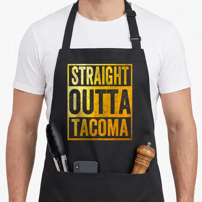 Outta tacoma apron - Stacked yellow and grey distressed text reading straight outta tacoma inside a yellow rectangular — shown on a black cotton canvas apron against a white background