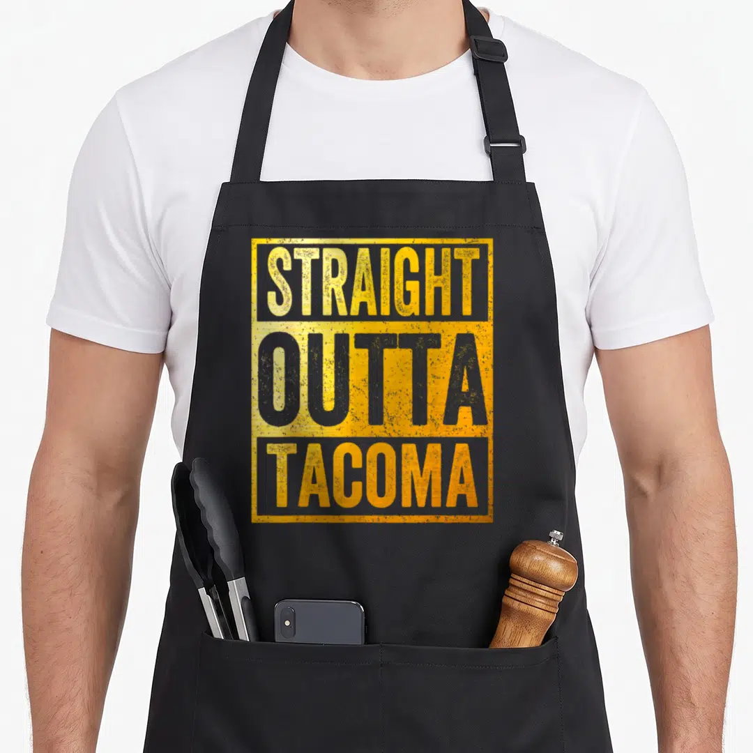 Outta tacoma apron - Stacked yellow and grey distressed text reading straight outta tacoma inside a yellow rectangular — shown on a black cotton canvas apron against a white background