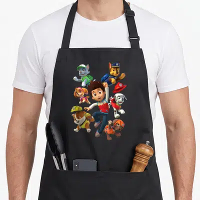Ryder novelty apron - Boy surrounded by five dogs in blue red yellow and green cartoon rescue gear — shown on a black cotton canvas apron against a white background