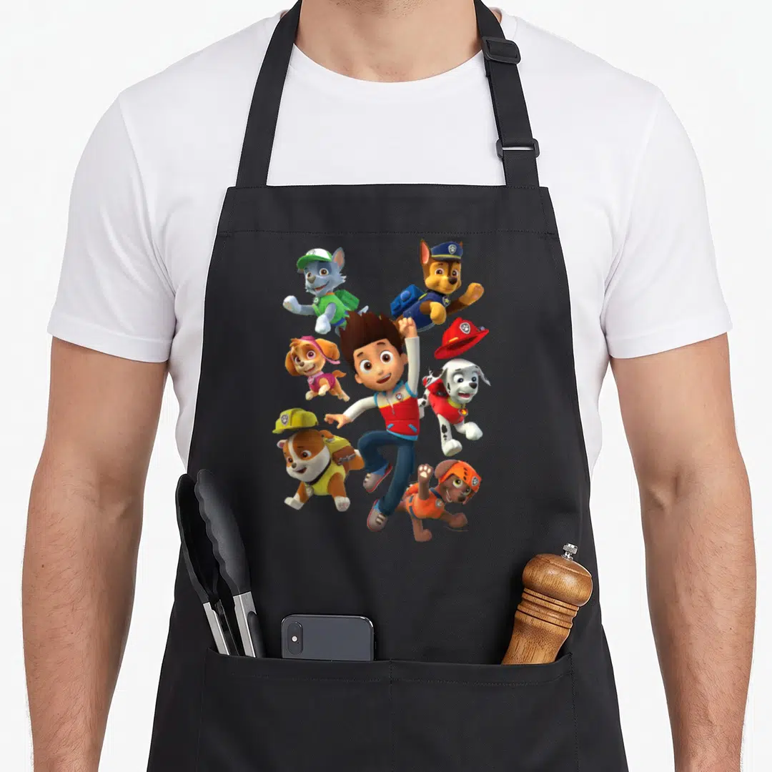 Ryder novelty apron - Boy surrounded by five dogs in blue red yellow and green cartoon rescue gear — shown on a black cotton canvas apron against a white background