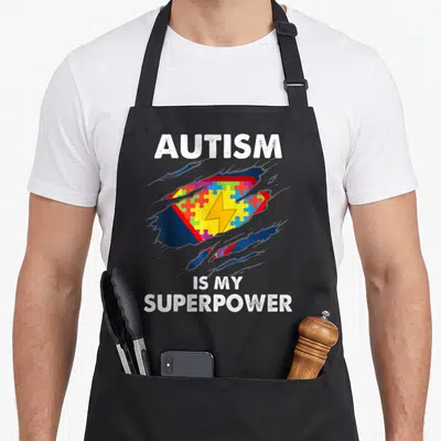 Autism superhero apron - Torn graphic revealing colorful puzzle piece shield with lightning bolt and white text reading — shown on a black cotton canvas apron against a white background