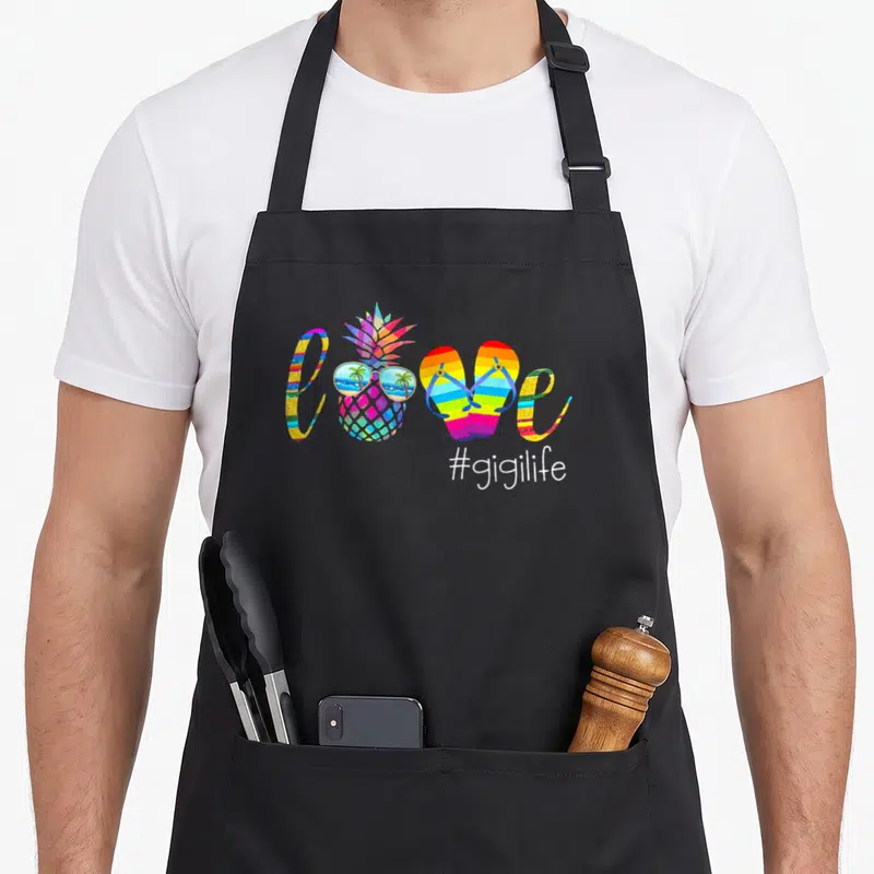 Gigi pineapple apron - Rainbow tie-dye love spelled with pineapple and flip flops above #gigilife text — shown on a black cotton canvas apron against a white background