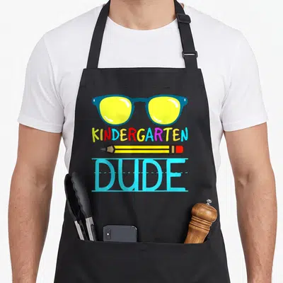 School dude apron - Glasses, multi-colored KINDERGARTEN text, pencil, and DUDE on handwriting lines — shown on a black cotton canvas apron against a white background