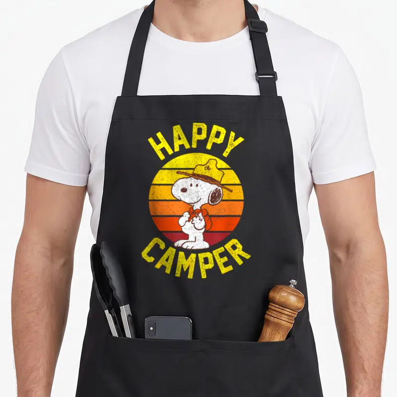 Cute happy camper apron - Snoopy wearing a hat in front of a striped yellow and orange sunset circle — shown on a black cotton canvas apron against a white background