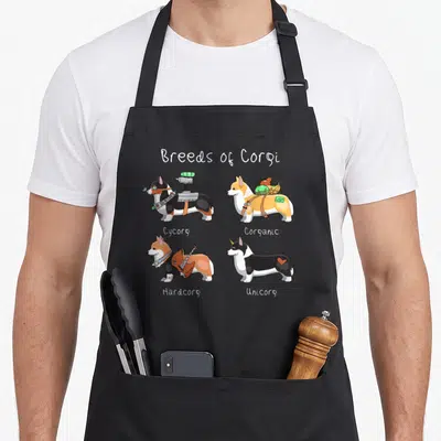 Breed corgi apron - Four cartoon corgis labeled Cycorg Corganic Hardcorg Unicorg with accessories — shown on a black cotton canvas apron against a white background