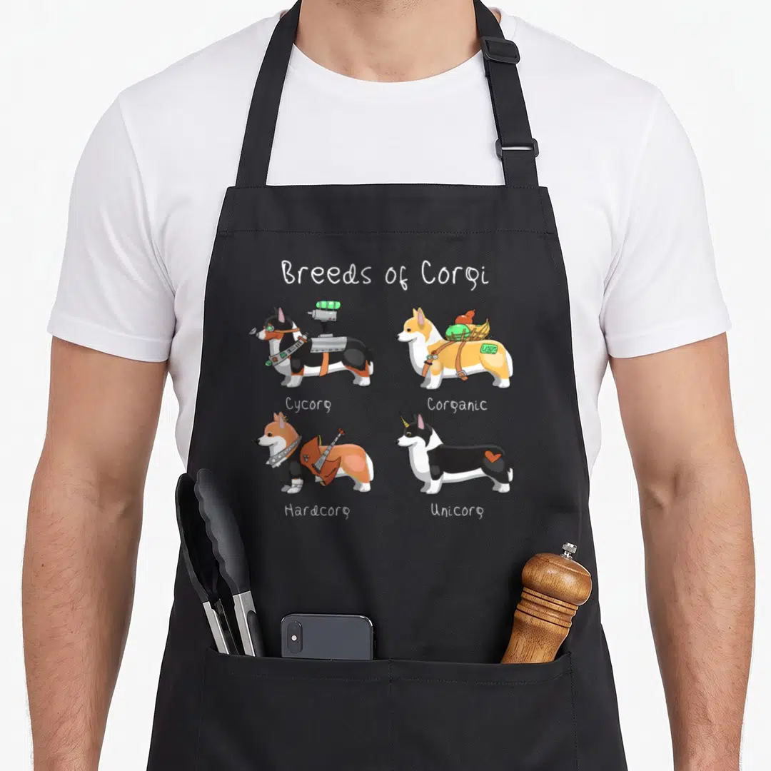 Breed corgi apron - Four cartoon corgis labeled Cycorg Corganic Hardcorg Unicorg with accessories — shown on a black cotton canvas apron against a white background