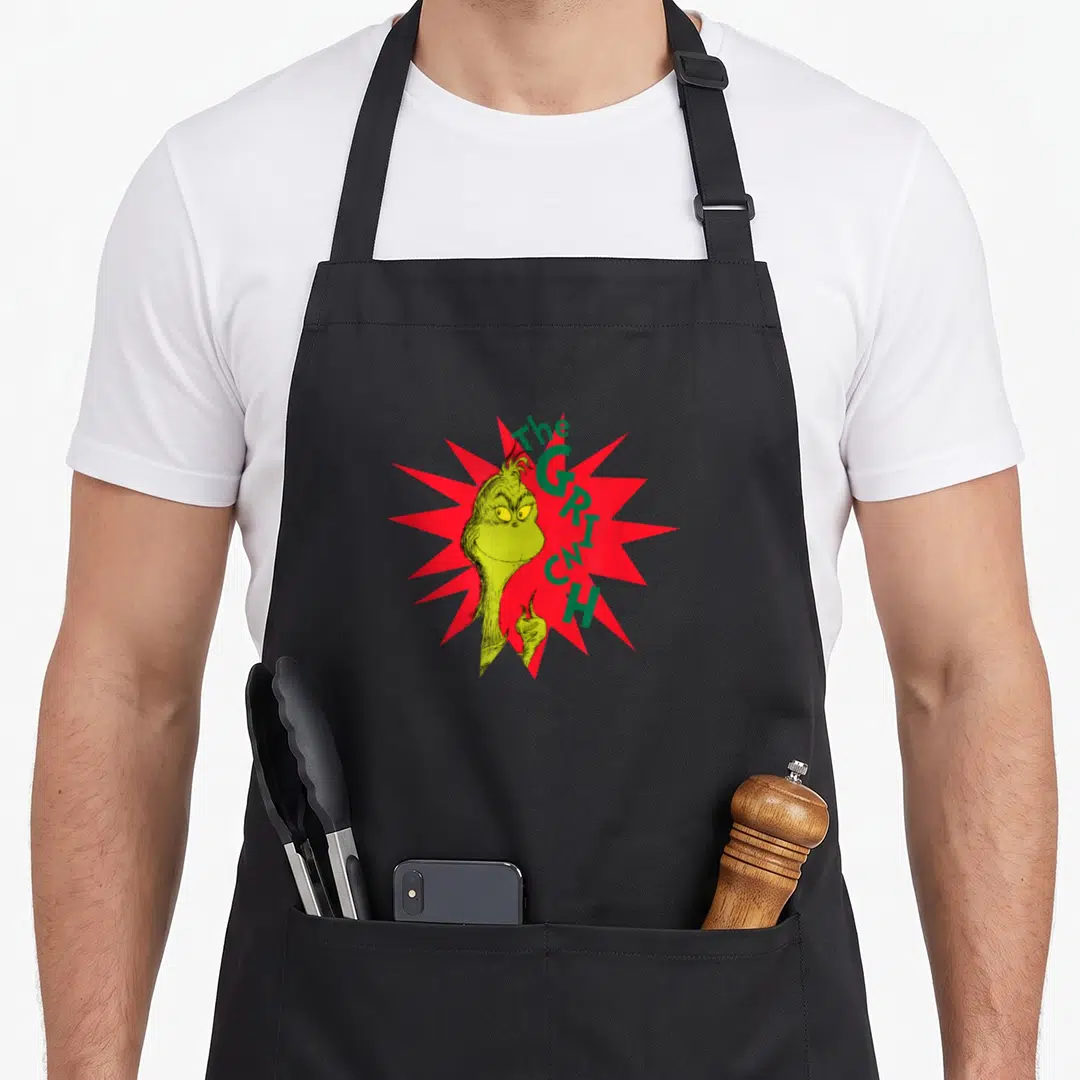 Burst holiday apron - Cartoon Grinch character in lime green with red starburst and curved text — shown on a black cotton canvas apron against a white background