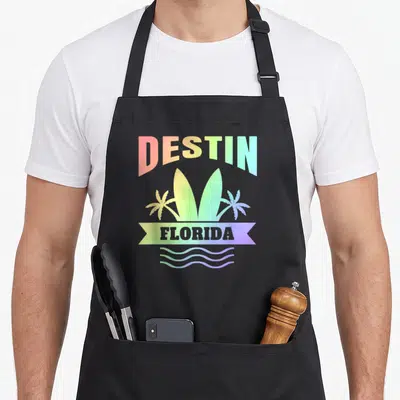 Destin florida family aprons - Arched DESTIN text above two surfboards, palm trees, FLORIDA banner, and three wavy — shown on a black cotton canvas apron against a white background