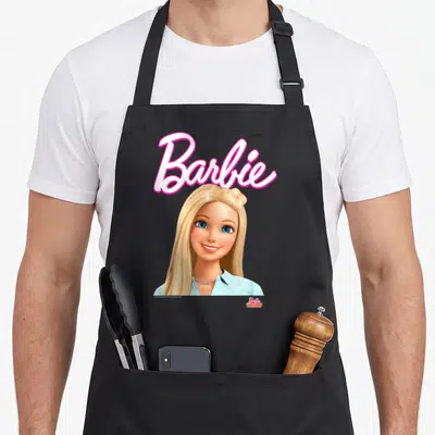 Dreamhouse little miss apron - Pink Barbie script above a blonde woman in a blue shirt with blue eyes — shown on a black cotton canvas apron against a white background
