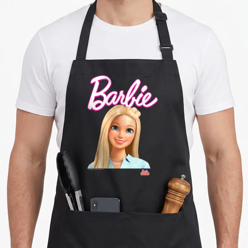 Dreamhouse little miss apron - Pink Barbie script above a blonde woman in a blue shirt with blue eyes — shown on a black cotton canvas apron against a white background