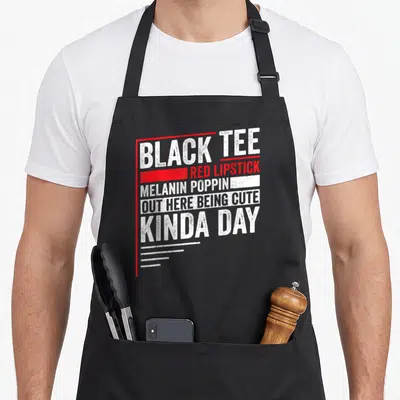 Lipstick novelty apron - Stacked white and red text: BLACK TEE RED LIPSTICK MELANIN POPPIN OUT HERE BEING CUTE KINDA — shown on a black cotton canvas apron against a white background