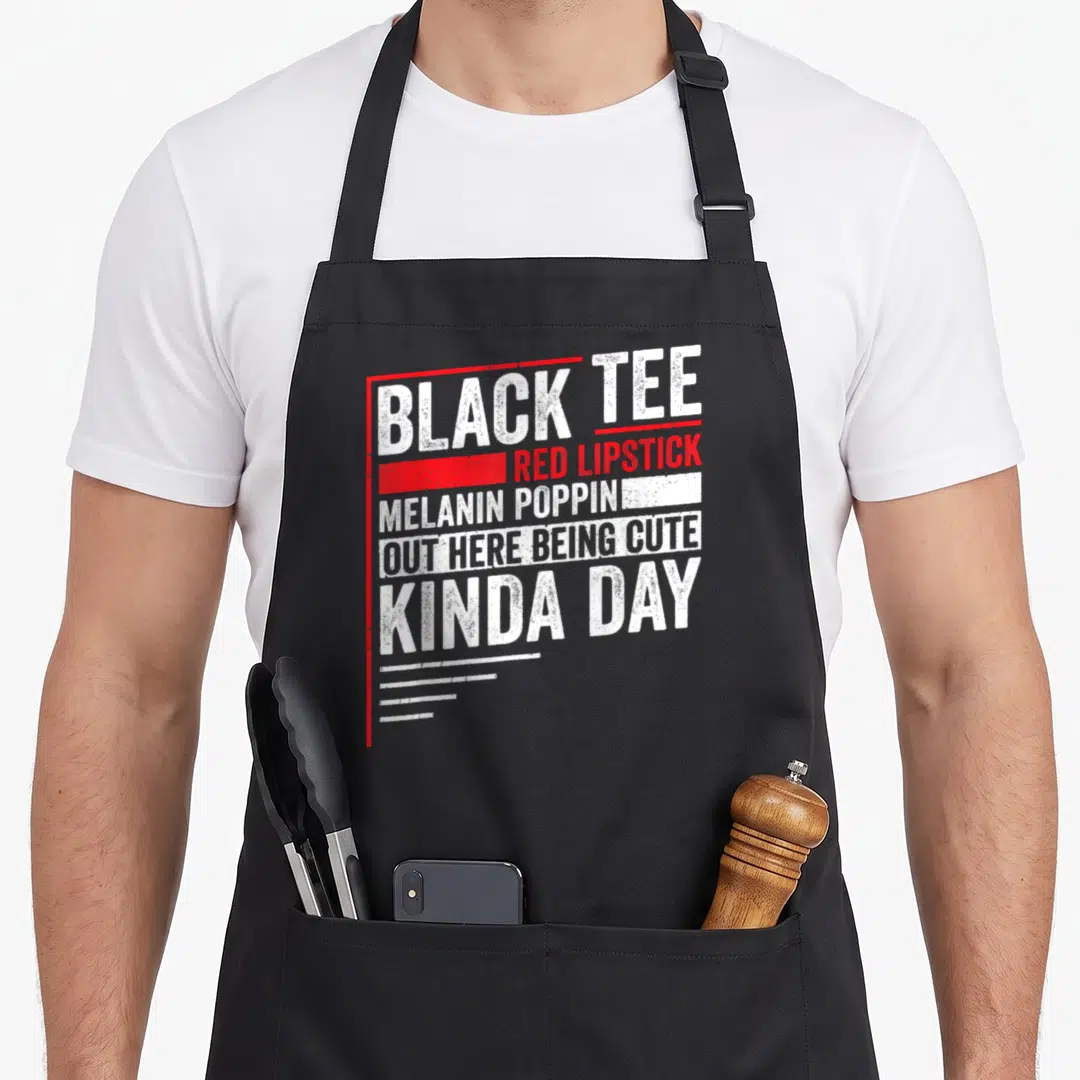 Lipstick novelty apron - Stacked white and red text: BLACK TEE RED LIPSTICK MELANIN POPPIN OUT HERE BEING CUTE KINDA — shown on a black cotton canvas apron against a white background