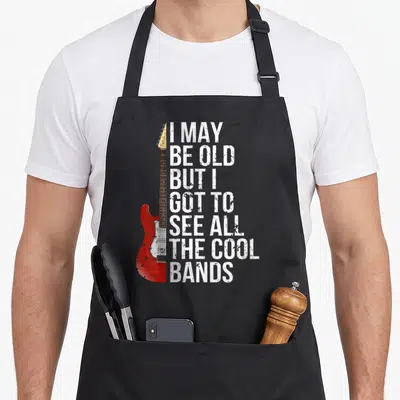 See may apron - Red electric guitar beside white distressed text I MAY BE OLD BUT I GOT TO SEE ALL THE COOL BANDS — shown on a black cotton canvas apron against a white background