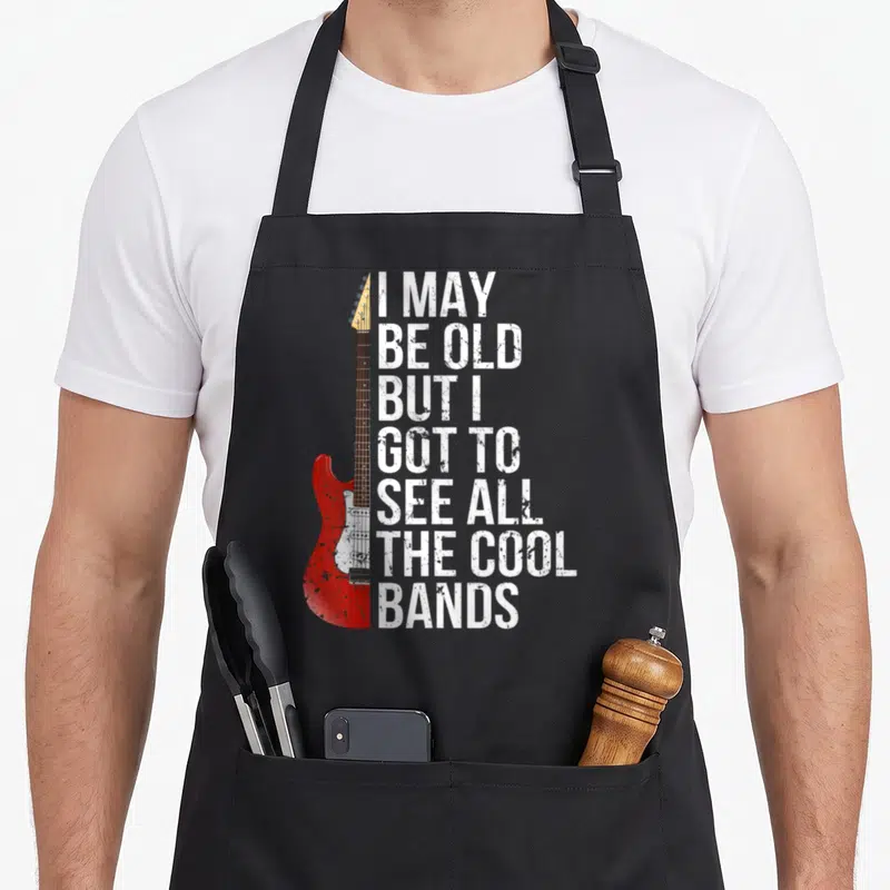 See may apron - Red electric guitar beside white distressed text I MAY BE OLD BUT I GOT TO SEE ALL THE COOL BANDS — shown on a black cotton canvas apron against a white background