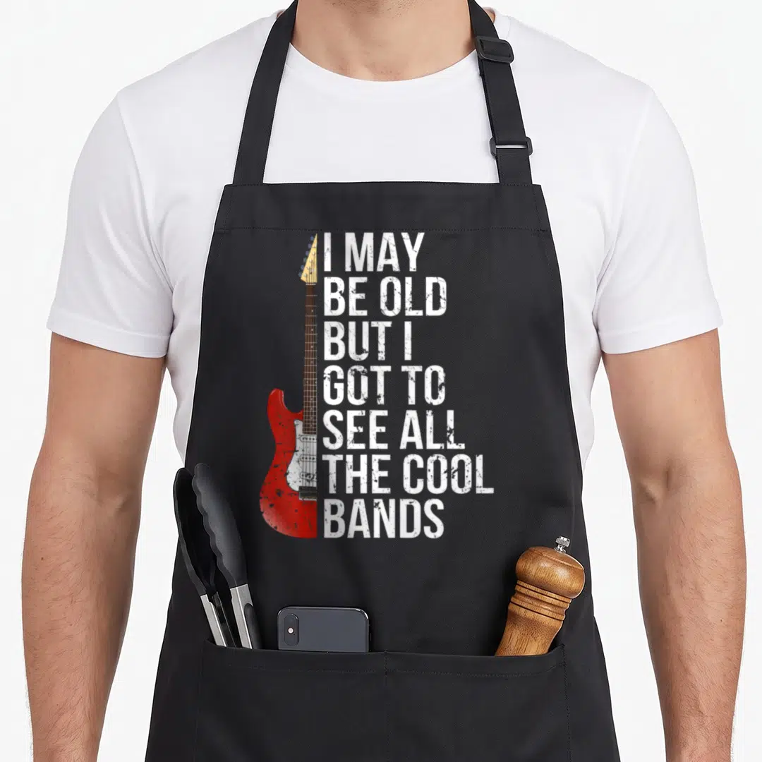See may apron - Red electric guitar beside white distressed text I MAY BE OLD BUT I GOT TO SEE ALL THE COOL BANDS — shown on a black cotton canvas apron against a white background