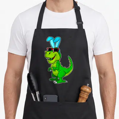 Rex basket apron - Green cartoon T-Rex wearing blue bunny ears and red sunglasses on a black background — shown on a black cotton canvas apron against a white background