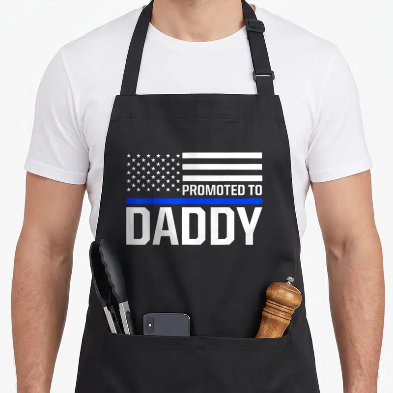 Police daddy apron - White flag graphic with Promoted To Daddy text and thin blue line — shown on a black cotton canvas apron against a white background
