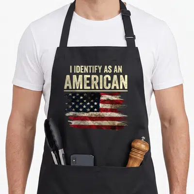 Identify 4th of july apron - Stacked beige text I IDENTIFY AS AN AMERICAN above distressed red white blue USA flag — shown on a black cotton canvas apron against a white background