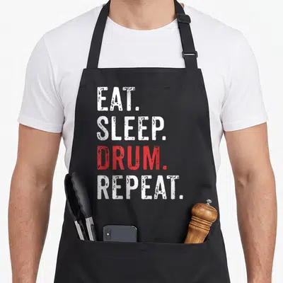 Eat drums apron - Stacked white and red text EAT SLEEP DRUM REPEAT on black fabric — shown on a black cotton canvas apron against a white background