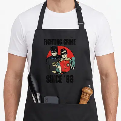 Cute tv apron - Retro Batman Robin illustration on red circle with fighting crime since 66 text — shown on a black cotton canvas apron against a white background