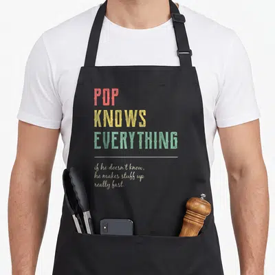 Funny pop grandpa apron - Stacked red yellow green text POP KNOWS EVERYTHING above white line and script disclaimer — shown on a black cotton canvas apron against a white background