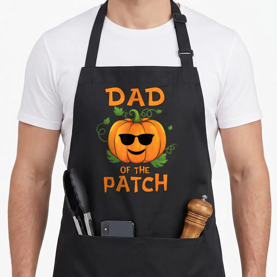 Pumpkin patch halloween apron - Cartoon orange pumpkin with sunglasses and DAD OF THE PATCH text surrounded by green — shown on a black cotton canvas apron against a white background