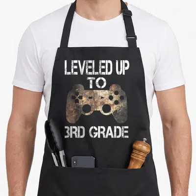 Leveled grade apron - Distressed game controller with stacked white text reading leveled up to 3rd grade — shown on a black cotton canvas apron against a white background