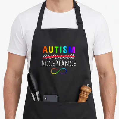 Instead autism apron - Rainbow AUTISM text with awareness crossed out and ACCEPTANCE above infinity symbol — shown on a black cotton canvas apron against a white background