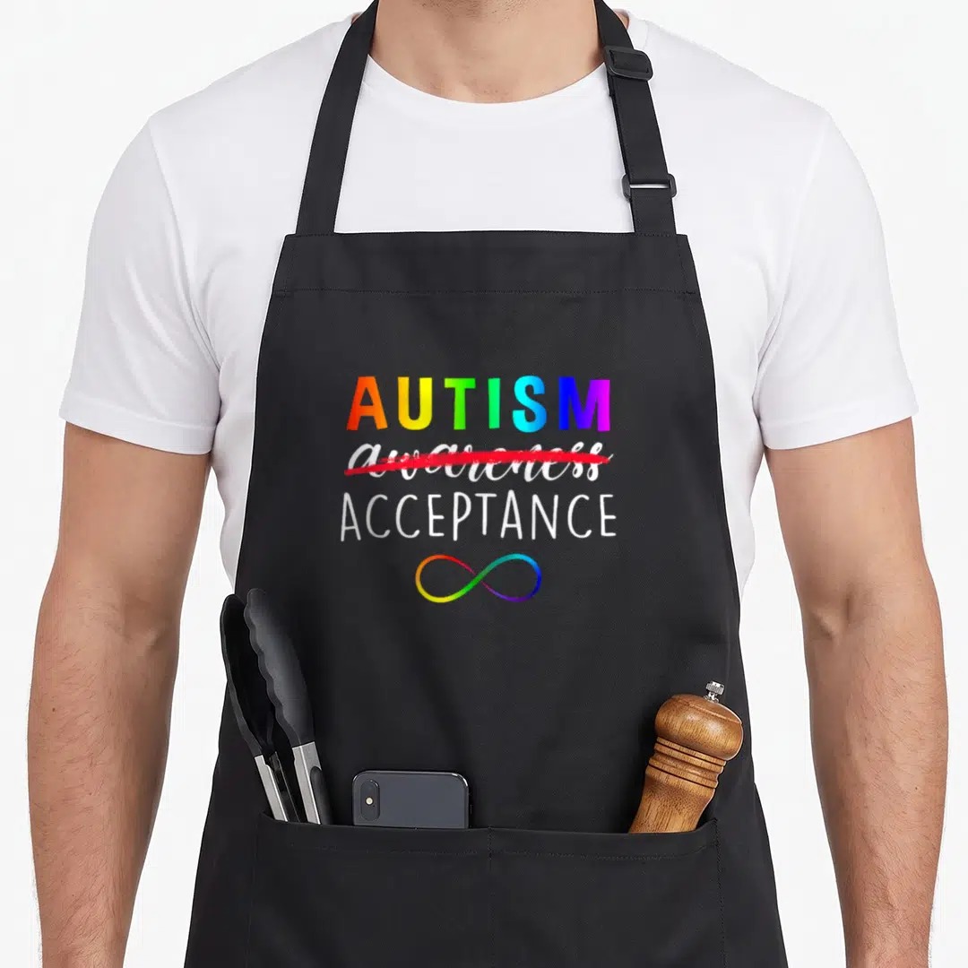 Instead autism apron - Rainbow AUTISM text with awareness crossed out and ACCEPTANCE above infinity symbol — shown on a black cotton canvas apron against a white background