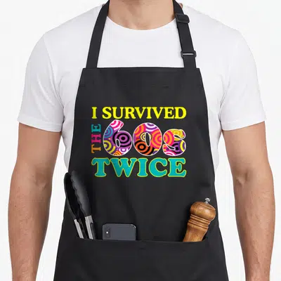Survived 60th birthday apron - Retro text I Survived The 60s Twice with colorful concentric circles — shown on a black cotton canvas apron against a white background