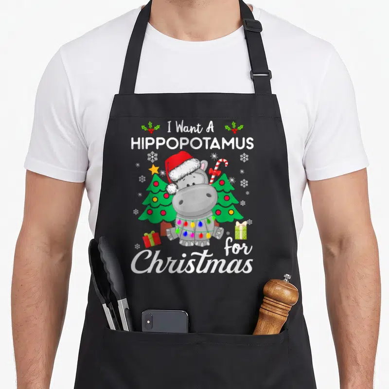 Costume christmas apron - Cartoon grey hippo in Santa hat holding candy cane between two Christmas trees with text I — shown on a black cotton canvas apron against a white background
