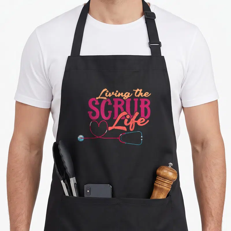 Living nurse apron - Stacked living scrub life text above a pink and blue stethoscope shaped into a heart — shown on a black cotton canvas apron against a white background