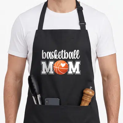 Funny basketball mother's apron - White basketball text above MOM with orange ball and white heart replacing the letter — shown on a black cotton canvas apron against a white background