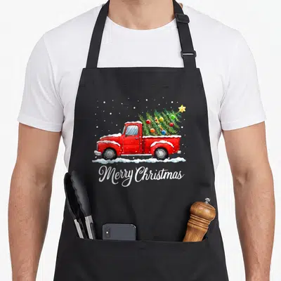 Merry old fashioned aprons - Red truck carrying decorated Christmas tree with Merry Christmas script in watercolor style — shown on a black cotton canvas apron against a white background