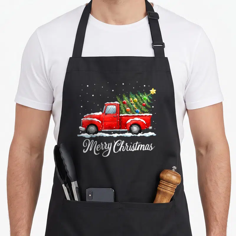 Merry old fashioned aprons - Red truck carrying decorated Christmas tree with Merry Christmas script in watercolor style — shown on a black cotton canvas apron against a white background