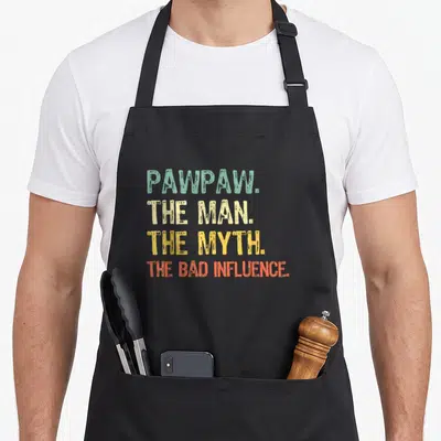 Myth papaya apron - Stacked retro text PAWPAW THE MAN THE MYTH THE BAD INFLUENCE in green beige yellow orange — shown on a black cotton canvas apron against a white background
