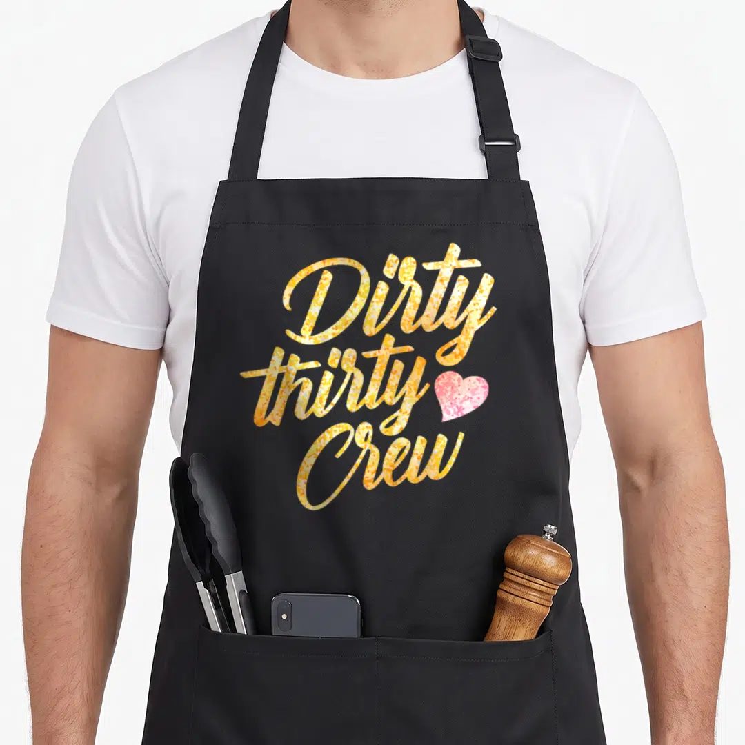 Thirty crew funny dirty aprons - Yellow and orange script text reading Dirty thirty Crew with a pink heart on grey — shown on a black cotton canvas apron against a white background