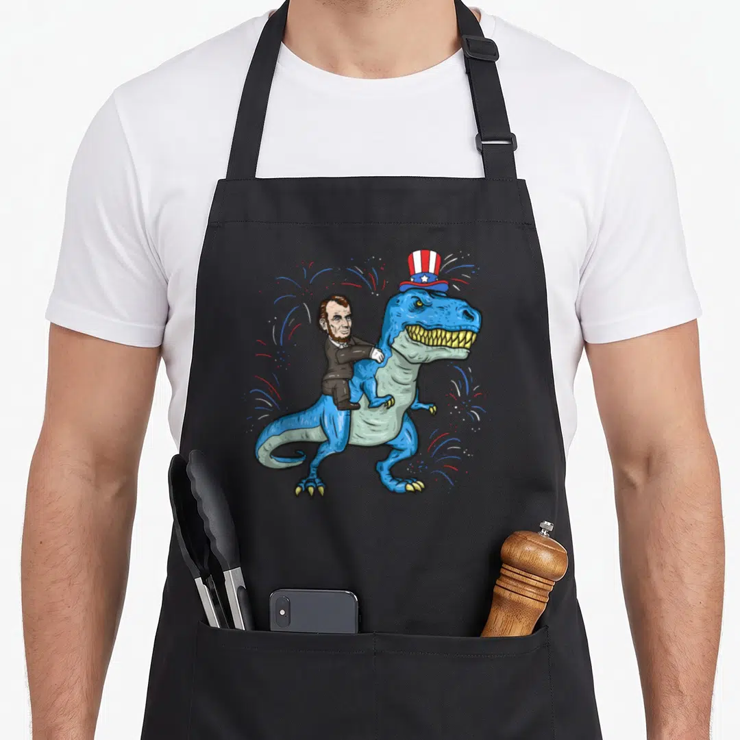 Abe riding 4th of july apron - Cartoon Abraham Lincoln riding a blue T-Rex wearing a patriotic top hat with red white — shown on a black cotton canvas apron against a white background