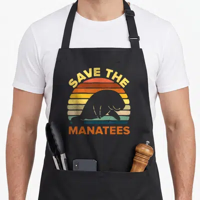 Save manatee apron - Grey manatee silhouette over striped orange and yellow sun with distressed SAVE THE MANATEES text — shown on a black cotton canvas apron against a white background
