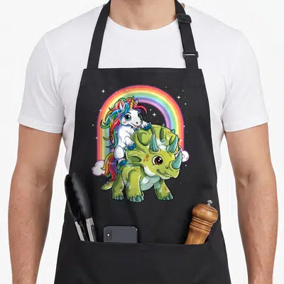 Unicorn riding dinosaur apron - White unicorn with rainbow mane sitting on green triceratops under rainbow arch — shown on a black cotton canvas apron against a white background