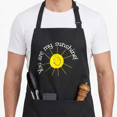 Sunshine apron or apron - Smiling yellow sun with white arched text You are my sunshine! — shown on a black cotton canvas apron against a white background