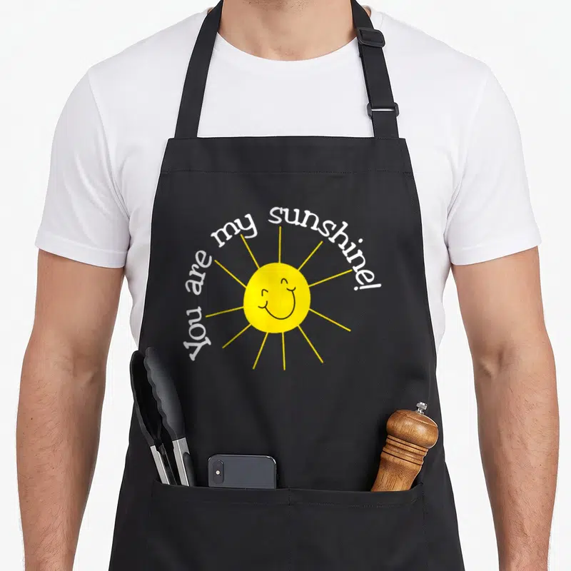 Sunshine apron or apron - Smiling yellow sun with white arched text You are my sunshine! — shown on a black cotton canvas apron against a white background