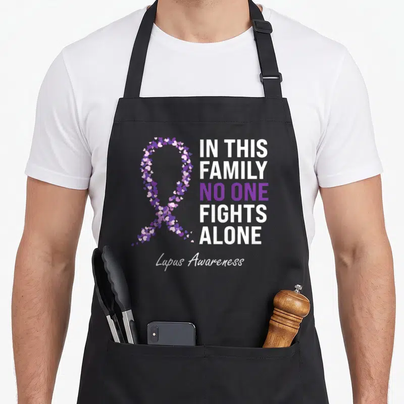 Funny lupus ribbon apron - Ribbon formed by small purple and white hearts next to stacked text reading IN THIS FAMILY — shown on a black cotton canvas apron against a white background