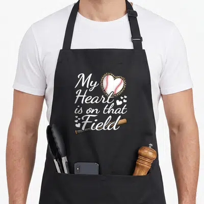 Field grandma apron - Baseball in leopard heart with My Heart is on that Field script and wooden bat — shown on a black cotton canvas apron against a white background