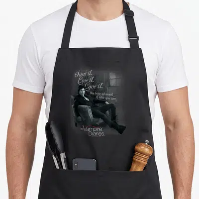 Yourself vampire apron - Man in armchair with script text Own it Live it Love it and Vampire Diaries logo — shown on a black cotton canvas apron against a white background